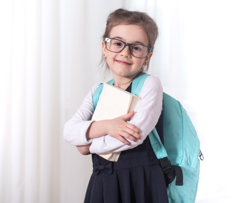 Girl-elementary school student with a backpack and a book with glasses is on a light background .The concept of education and primary school